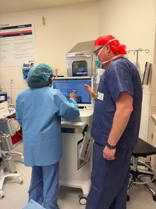Danny Minton in navy surgical scrubs and a red cap showing a colleague a touchscreen on an imaging cart in an operating room